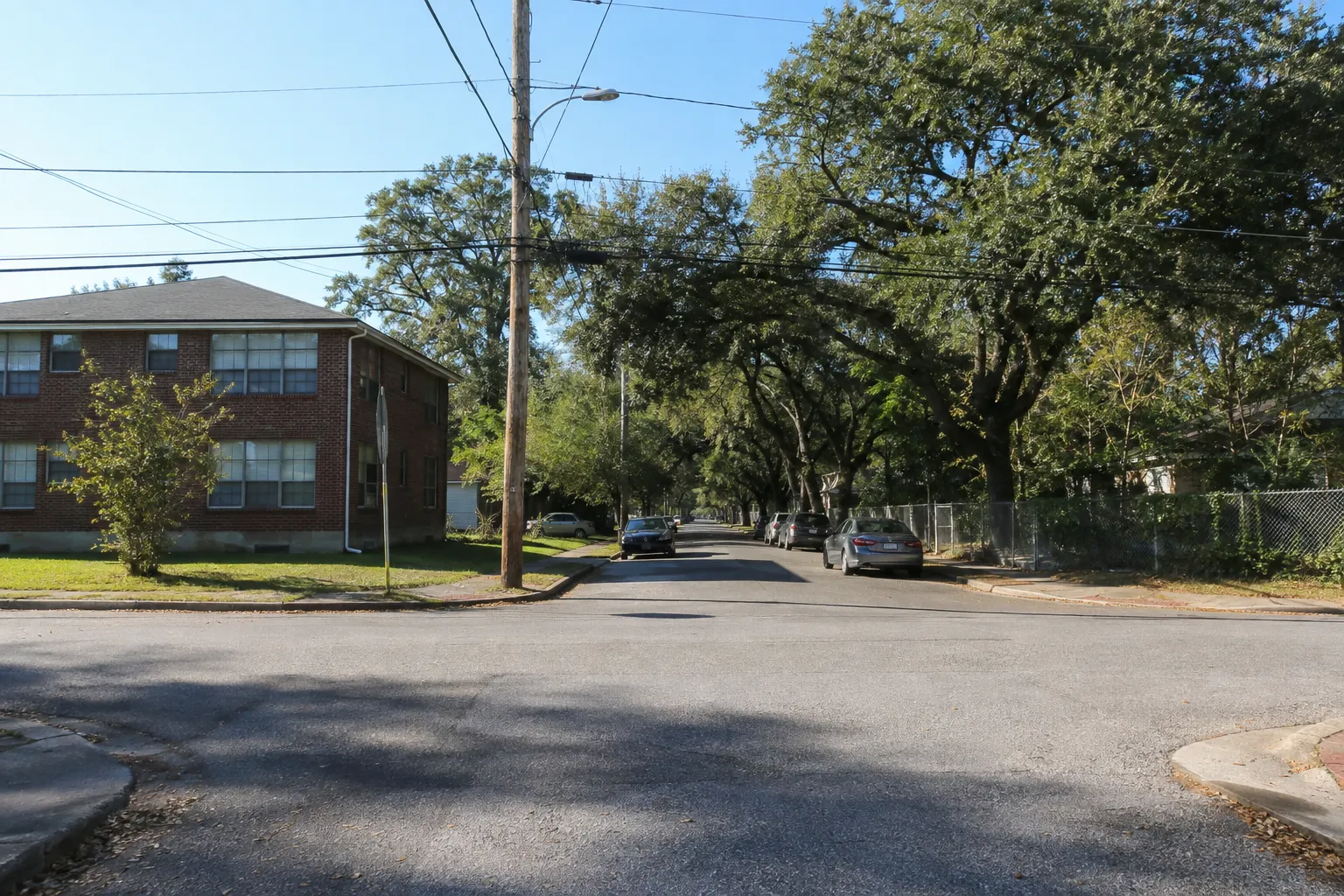 A residential street corner with a brick apartment building, parked cars, power lines and mature oaks, looking toward a tree-lined corridor a block away.